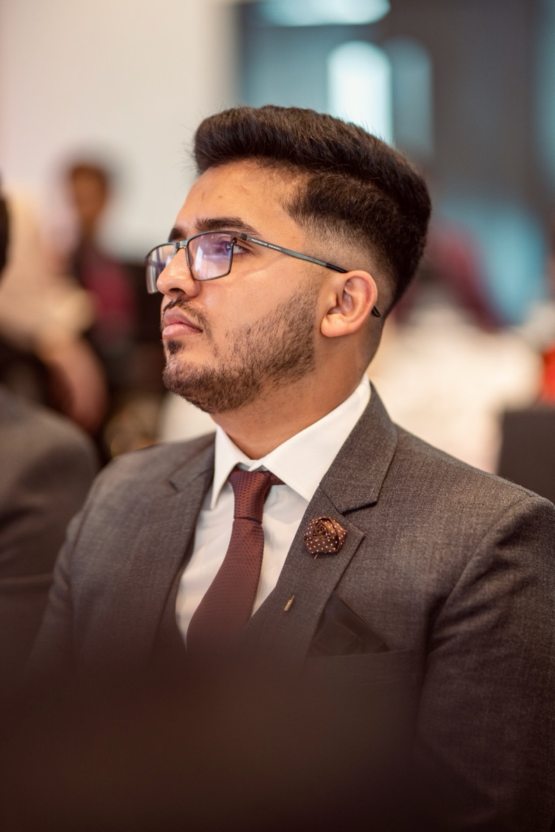 Muhammad Hamza seated at a professional event in a grey suit and burgundy tie